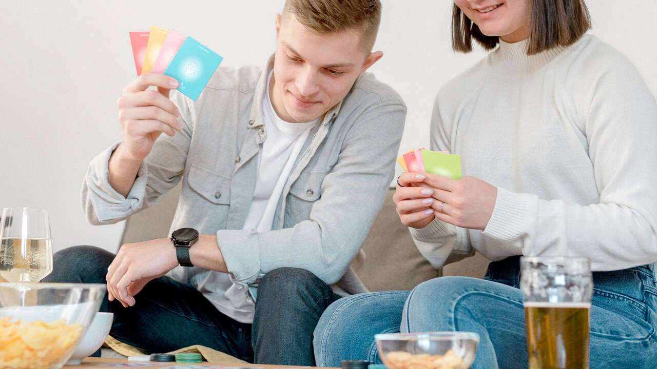 Hombre y mujer sosteniendo cartas de 'Encartados', un juego de cartas diseñado para actividad en pareja. Sentados en un ambiente informal con bebidas y aperitivos.