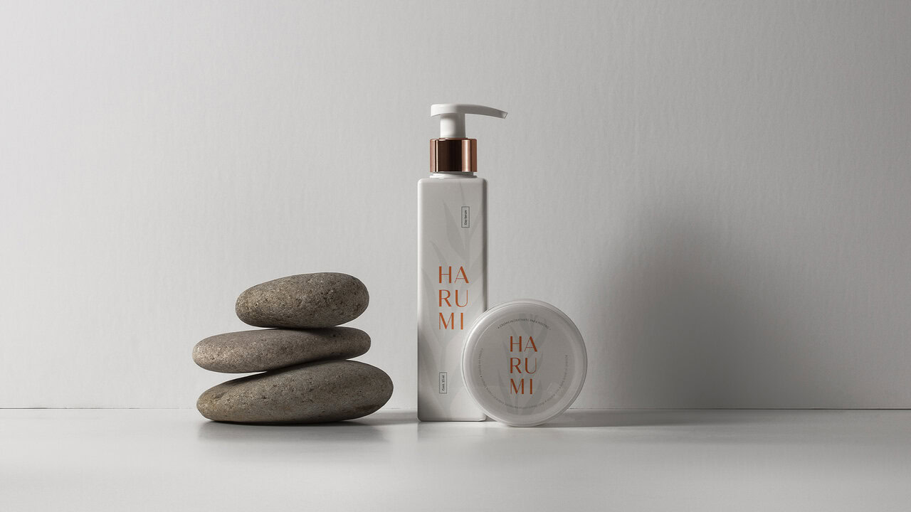 Varios productos Harumi (botella dispensadora y envase circular) junto a piedras apiladas. El diseño de empaque es minimalista y consistente con la identidad visual de la marca.