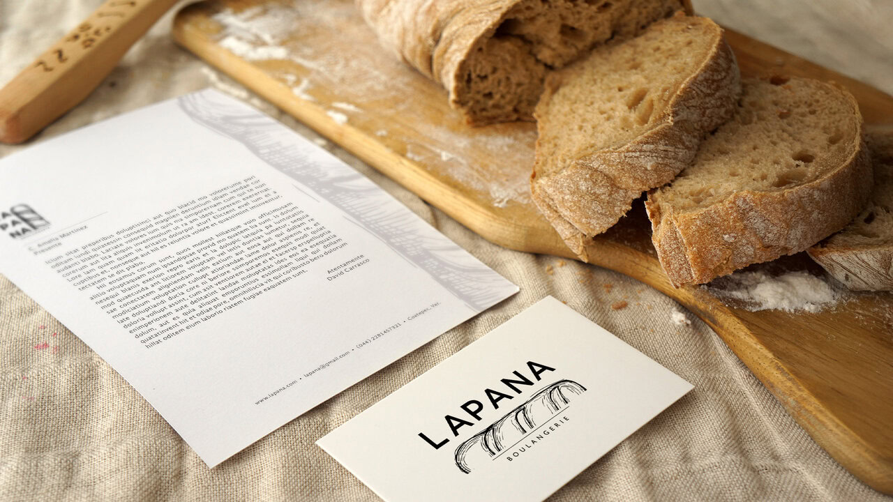 Elementos de papelería corporativa de LAPANA (tarjeta de presentación, hoja membretada) junto a un pan artesanal. Diseño de identidad para panadería artesanal.