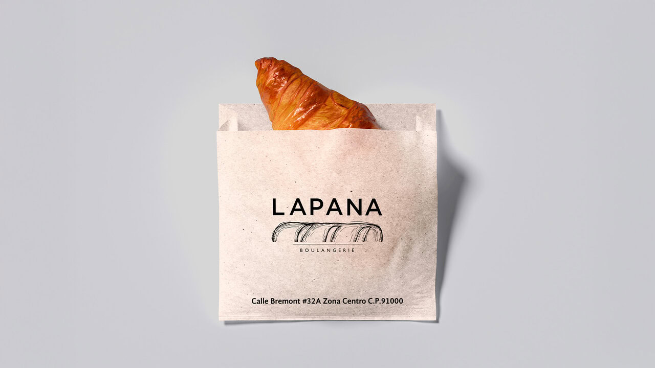 Bolsa de papel de con un croissant asomando. El diseño de empaque incluye el logotipo de LAPANA y su dirección. Branding acogedor para panadería artesanal: identidad cálida y tradición europea.