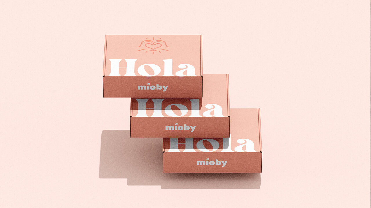 Cajas de envío de Mioby, apiladas. El diseño de empaque es en color rosa claro con el texto Hola y un logotipo de manos formando un corazón.