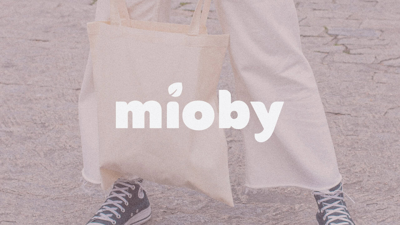 Persona de pie, vistiendo pantalones blancos y zapatillas, sosteniendo una tote bag con el logotipo de la marca impreso en la bolsa. Identidad única para emprendedores: piezas hechas a mano y esencia cálida.