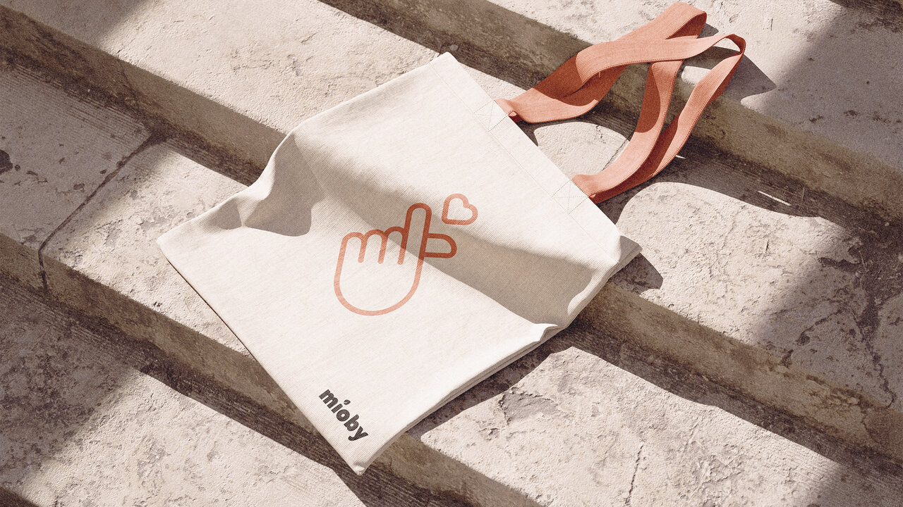 Tote bag de Mioby sobre escalones. Diseño en color crudo con el logotipo y una ilustración de una mano formando un corazón.