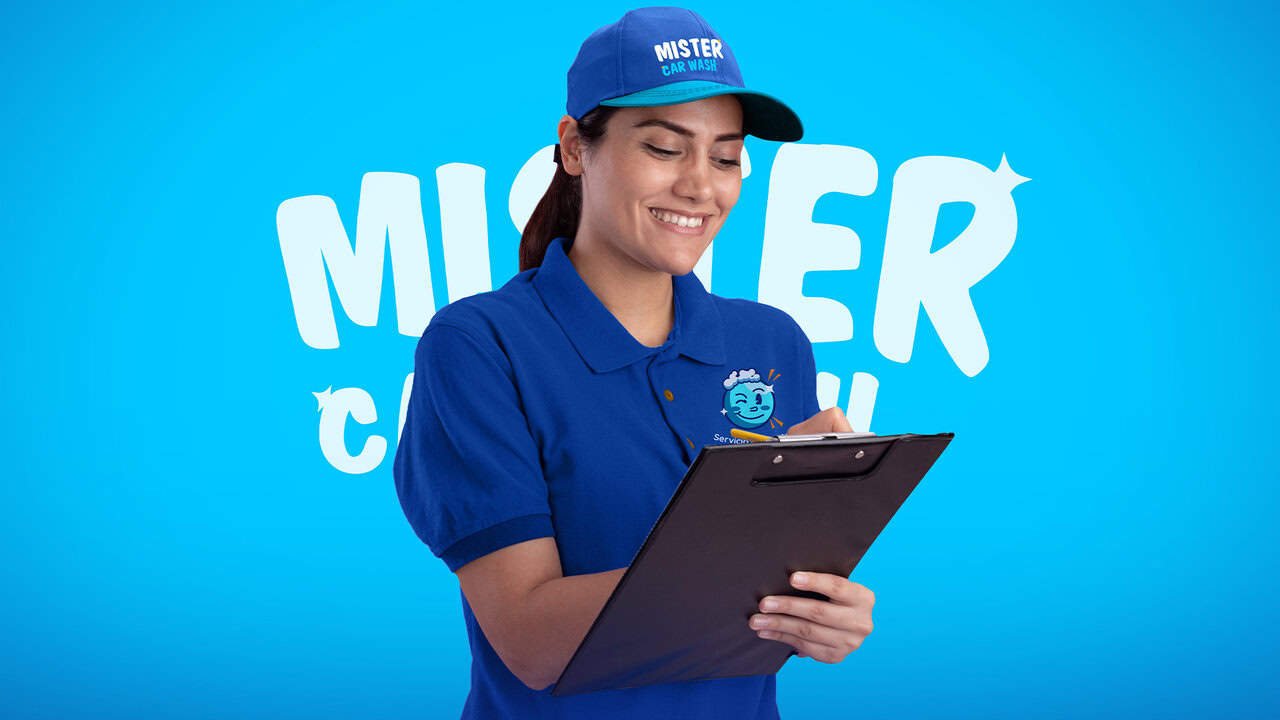 Mujer joven sonriente con una playera y gorra azul con el logo de mister car wash mientras escribe algo en un tablero.