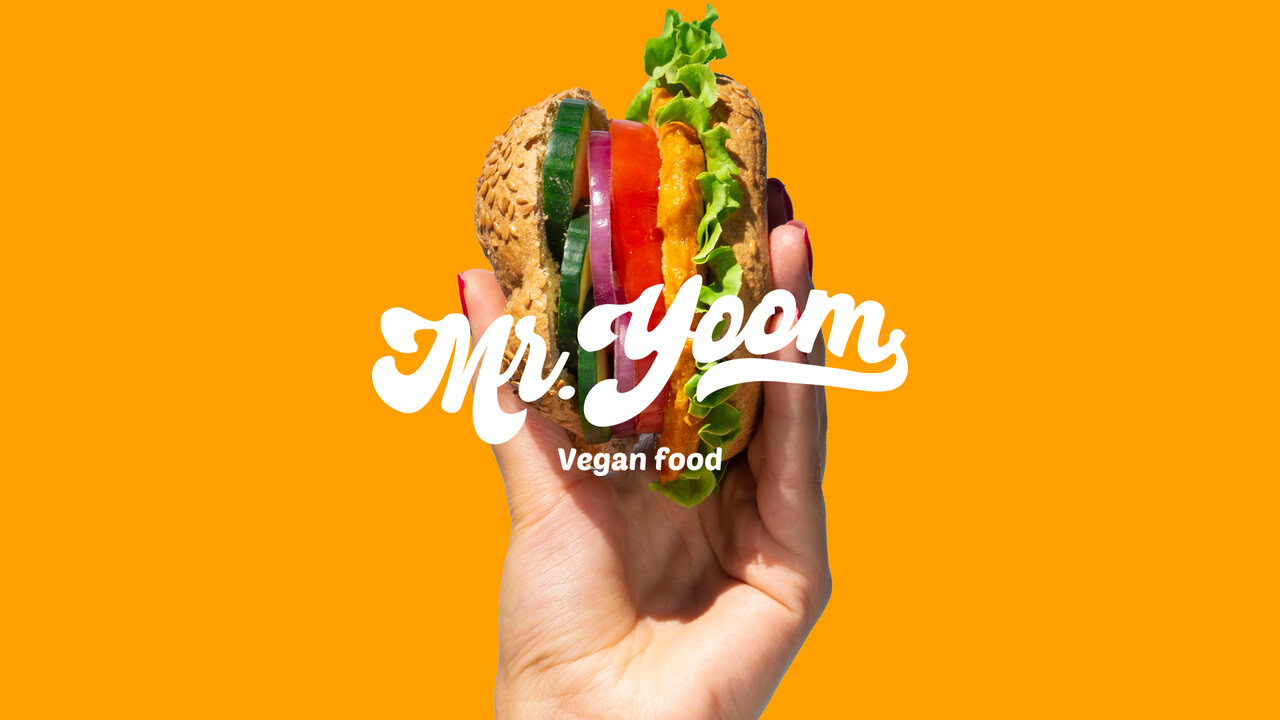 Mano sosteniendo una hamburguesa vegana con el logotipo de Mr. Yoom y el texto 'Vegan food' superpuestos. Fondo de color naranja sólido. Diseño publicitario para comida rápida vegana.