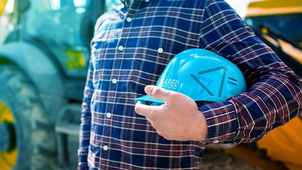 Persona con camisa a cuadros sosteniendo un casco de construcción azul con el logotipo de Gared. Al fondo, maquinaria de construcción. Identidad minimalista para despacho de construcción: enfoque innovador y moderno.